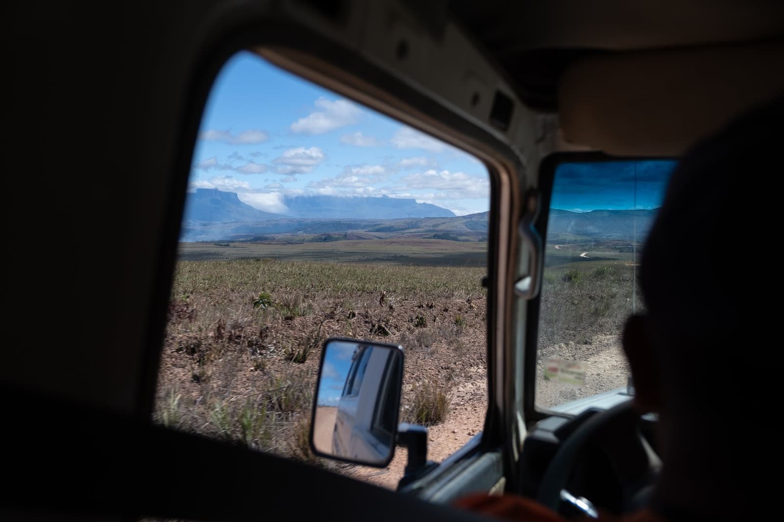 De tepui vult het raam al voordat je uit de truck stapt. Uitzicht door het zijraam van een 4x4-voertuig tijdens een Mount Roraima-expeditie, met een uitgestrekte savanne met lage struiken en een tafelbergachtige tepui aan de horizon onder een blauwe hemel met verspreide wolken
