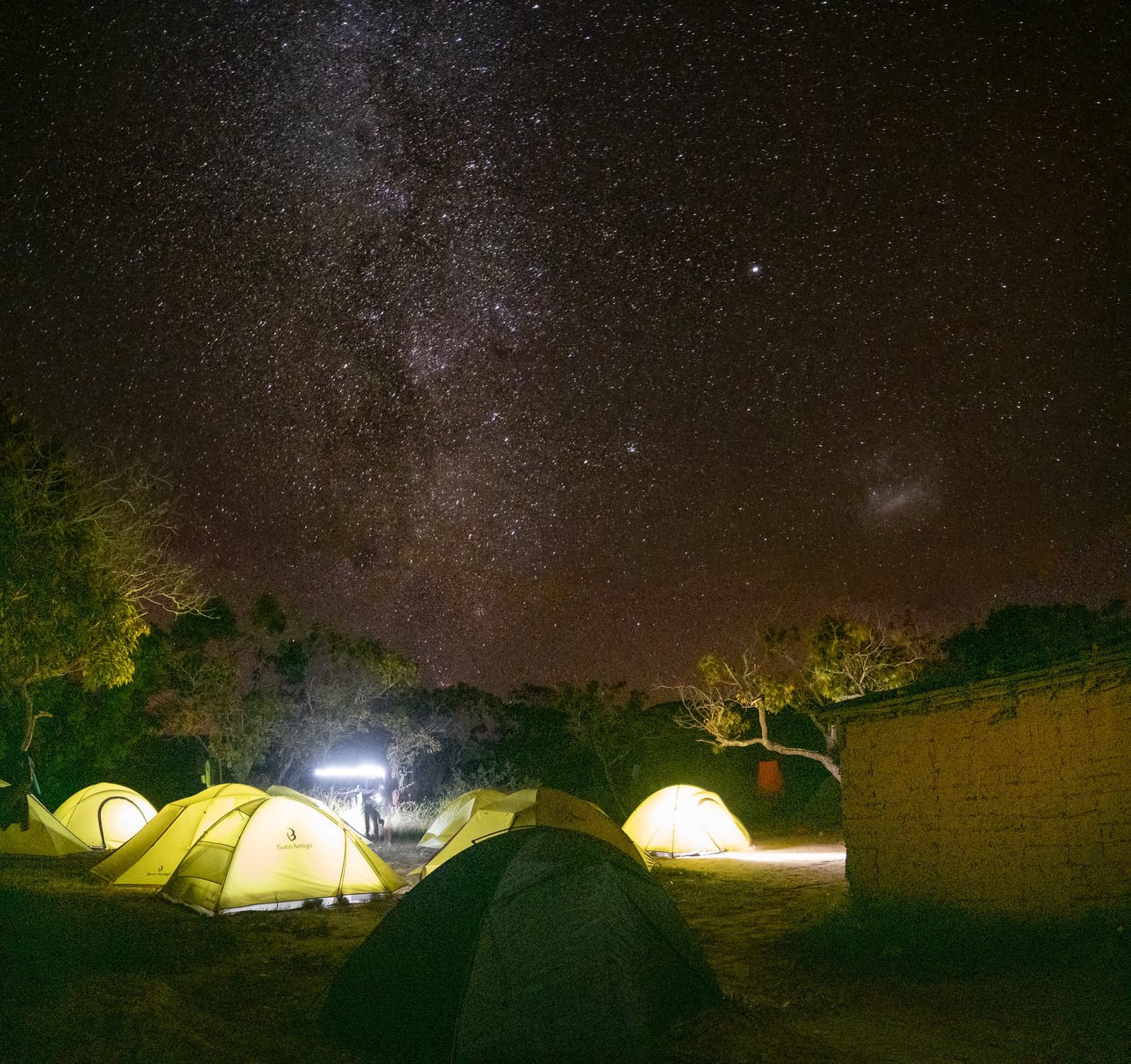 Zo ver van alles dat de Melkweg je buurman wordt. Verlichte gele koepeltenten op een kampeerplaats tijdens een Mount Roraima-expeditie, met de Melkweg die zich uitstrekt over een sterrenhemel boven schaarse bomen en een lemen muur
