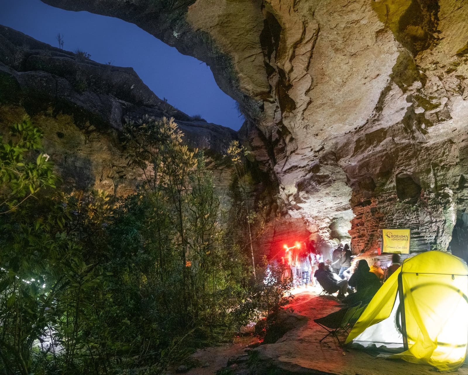 Een stenen dak, warm licht en goed gezelschap. Camp Coati maakt zijn naam waar. Een gloeiende gele tent opgezet onder een massief rotsoverhang tijdens een Mount Roraima-expeditie, met een groep trekkers verzameld rond rood lantaarnlicht diep in de grot-achtige schuilplaats 's nachts