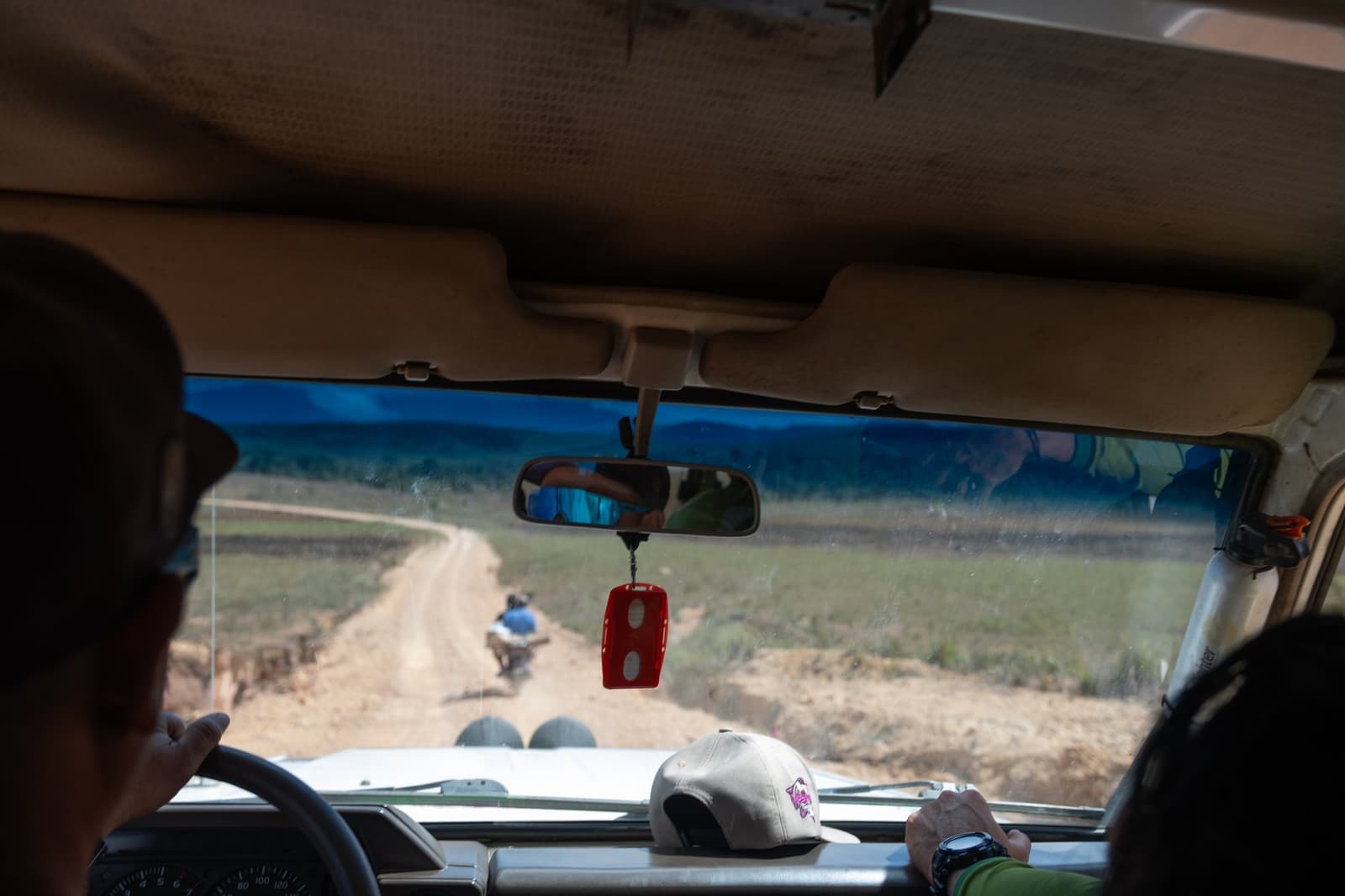 Onverharde weg, een bungelend sleutelhangertje, en de berg ergens verderop. Uitzicht vanuit een terreinwagen tijdens een Mount Roraima-expeditie, door de voorruit kijkend op een kronkelend onverhard pad door open grasland, met een motorfiets zichtbaar voor het voertuig en passagiers weerspiegeld in de achteruitkijkspiegel