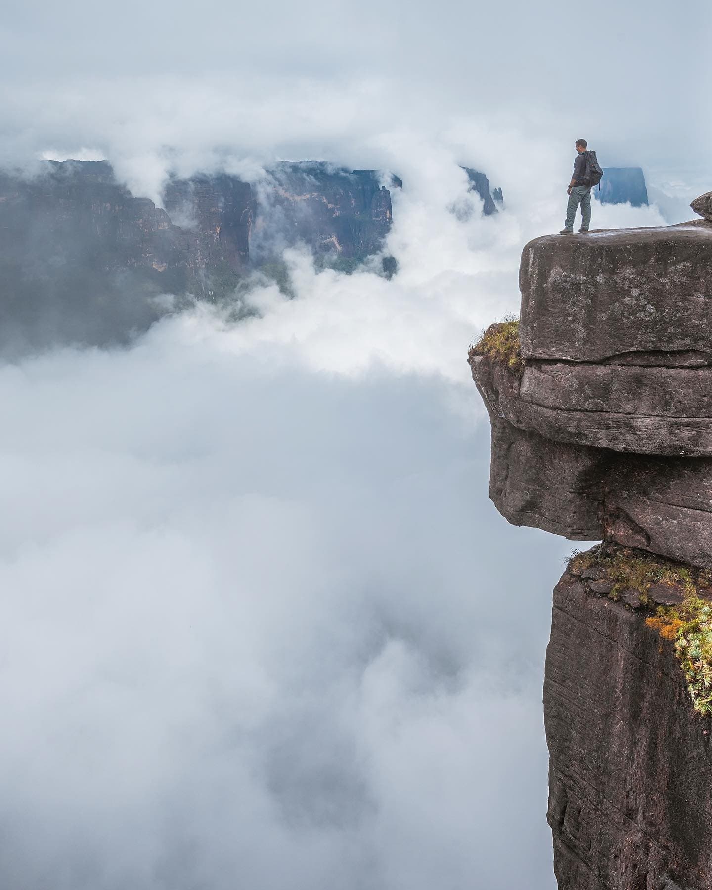 Op wolkenhoogte staan is hier geen uitdrukking. Het is gewoon de realiteit. Een rugzakreiziger staat alleen op een smal, eeuwenoud rotsuitsteeksel tijdens een Mount Roraima-expeditie, omgeven door een zee van wolken met een verre tepui-wand die door de mist opdoemt