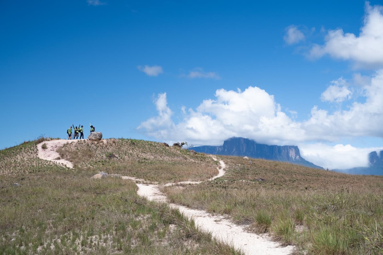 Eén iemand steekt zijn armen omhoog, en plots doet iedereen het mee. Vier wandelaars vieren feest op een zanderige heuvel tijdens de Mount Roraima-expeditie, met de tafelberg zichtbaar door de wolken op de achtergrond