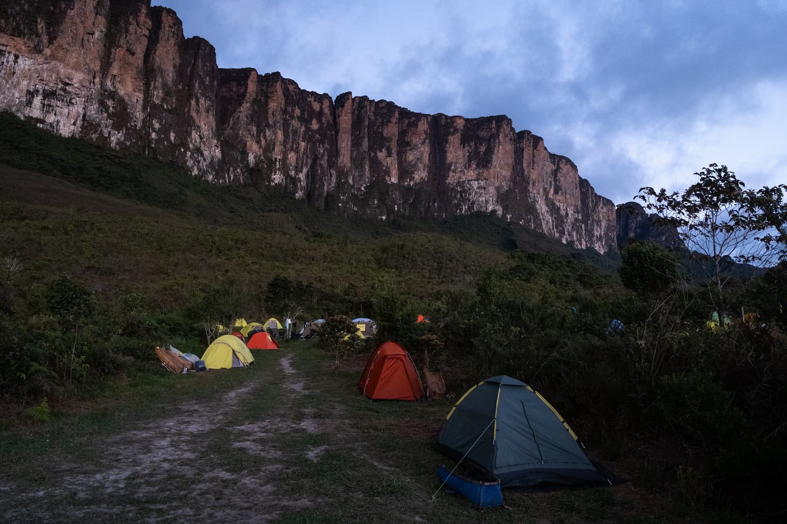 Tenten opgezet, rotswand vlakbij, morgen begint de klim. Kleurrijke koepeltenten opgesteld bij schemering op een grashelling tijdens een Mount Roraima-expeditie, met de imposante zandsteen rotswand die rood oplicht onder een bewolkte lucht