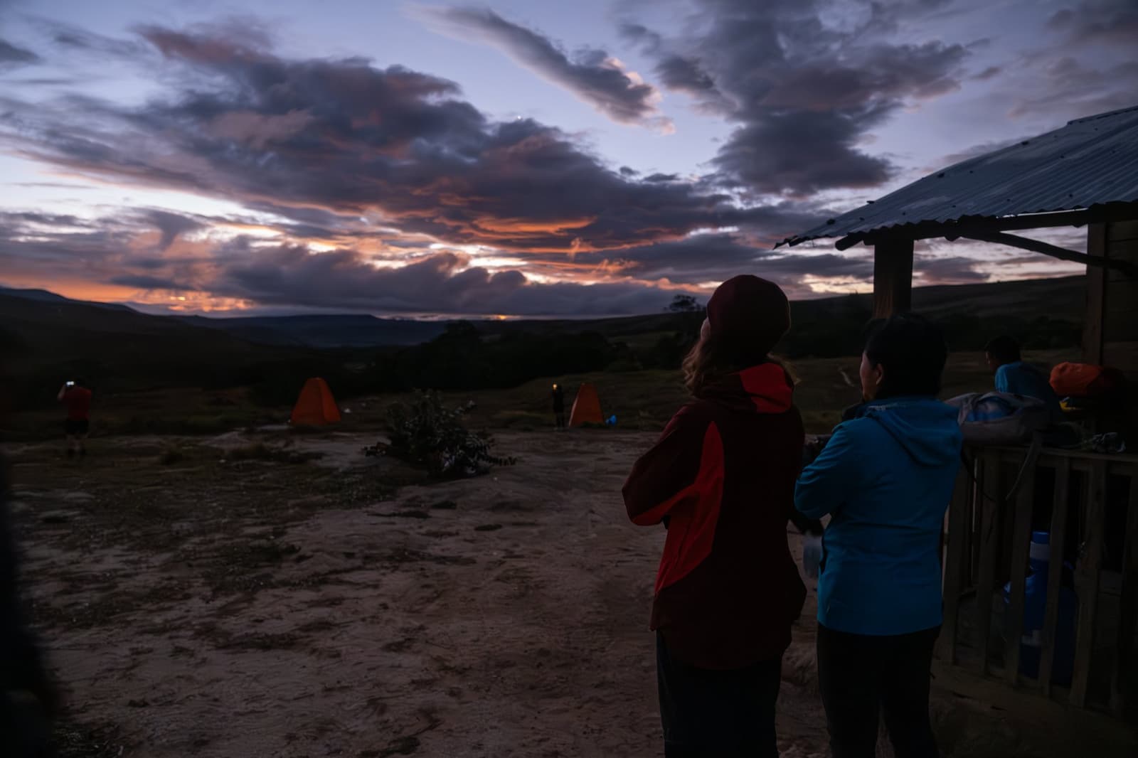 De zonsondergang wacht op niemand, maar vanavond lijkt hij een uitzondering te maken. Twee trekkers in warme jassen kijken naar een oranje-paarse zonsondergang vanuit een hooggelegen kampement tijdens een Mount Roraima-expeditie, met felgekleurde oranje tenten op de achtergrond
