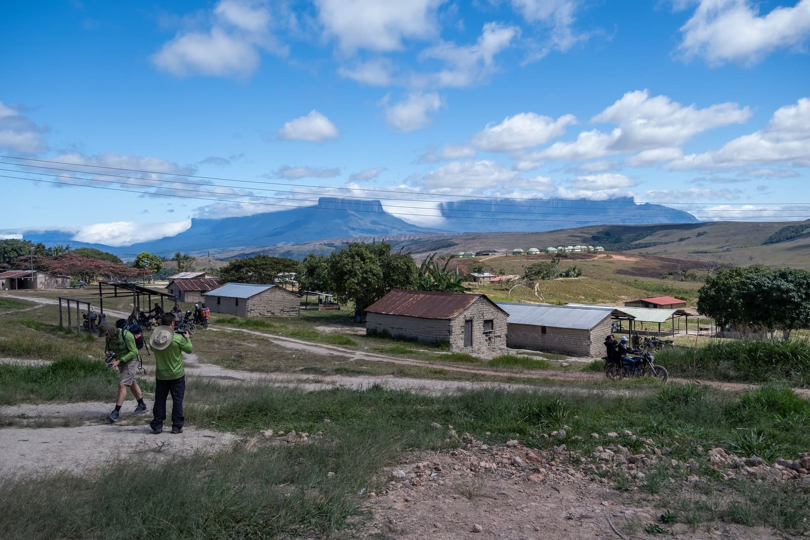 Paraitepuy: het laatste dorp voordat de berg het overneemt. Twee fotografen met camera's fotograferen het dorp Paraitepuy tijdens een Mount Roraima-expeditie, met stenen huizen, een onverharde weg en een tafelbergtepui die zich aftekenT aan de horizon onder een blauwe hemel