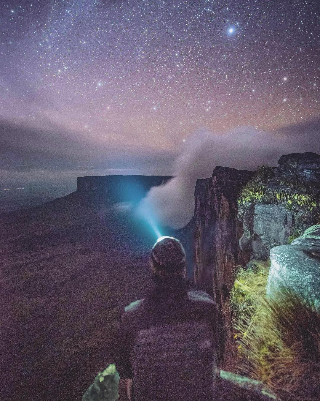 Soms geeft de berg je de hele hemel en vraagt hij niets terug. Een wandelaar met hoofdlamp staat aan de rand van een tepui-klif tijdens een Mount Roraima-expeditie en kijkt over een zee van mist naar een verre tafelberg onder een sterrenhemel