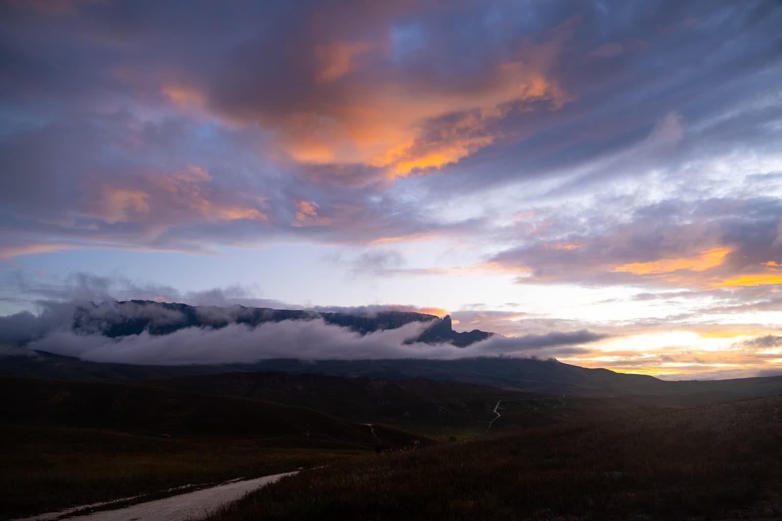 Laatste licht van de reis, en de berg heeft nog iets te zeggen. Groothoekzicht op een tafelberg die zich afteekent tegen een oranje-violette zonsonderganghemel tijdens een Mount Roraima-expeditie, met lage wolken die de klifrand omhullen en een onverharde weg die door donkere open savanne slingert op de voorgrond