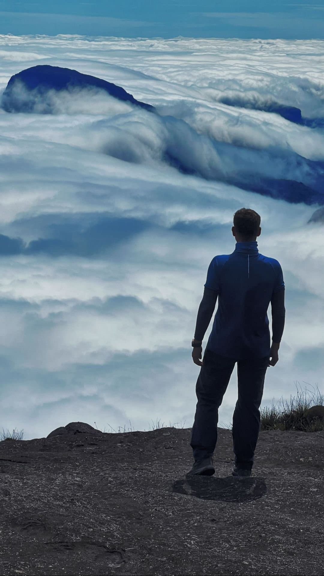 Boven de wolken uit, en de stilte neemt het over. Een wandelaar staat op een rotsrichel tijdens de Mount Roraima-expeditie en kijkt uit over een uitgestrekte wolkenzee waaruit donkere tepui-toppen nauwelijks zichtbaar zijn