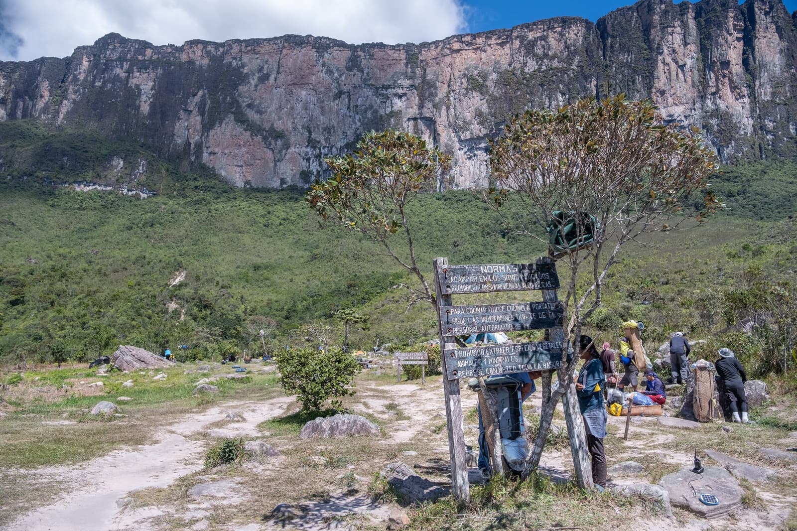 Regels gelezen, pakketten klaar, de rotswand kijkt al mee. Een houten regelbord met de tekst 'Normas' staat bij het basiskamp van een Mount Roraima-expeditie, terwijl dragers traditionele rugzakframes beladen en de loodrechte tepui-rotswand achter het kamp oprijst