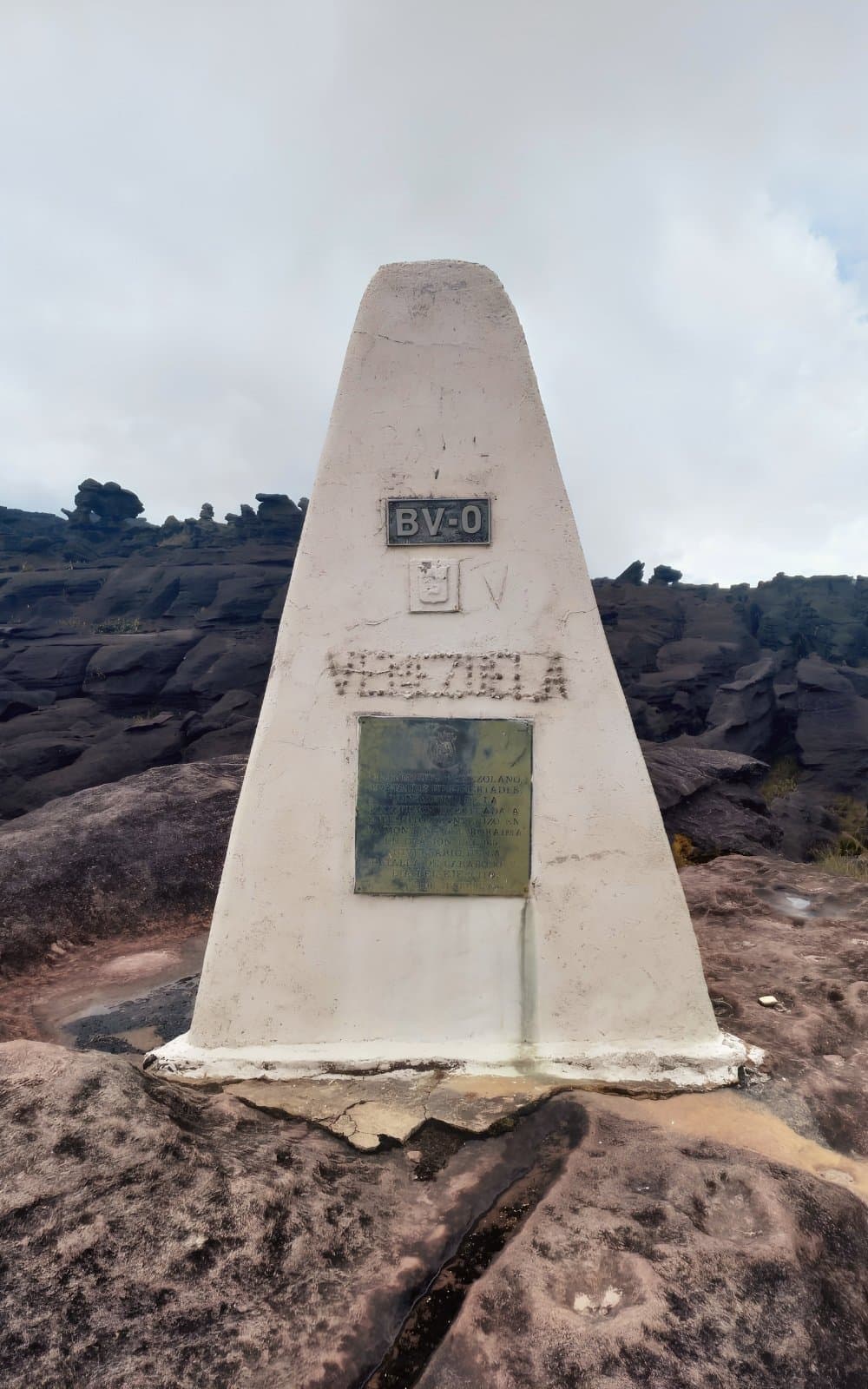 Drie landen, één betonnen obelisk, en een flinke tocht om er te komen. Witte piramidevormige grenssteen met de tekst BV-0 en Venezuela op donker oeroud gesteente op het drielandenpunt tijdens de Mount Roraima-expeditie