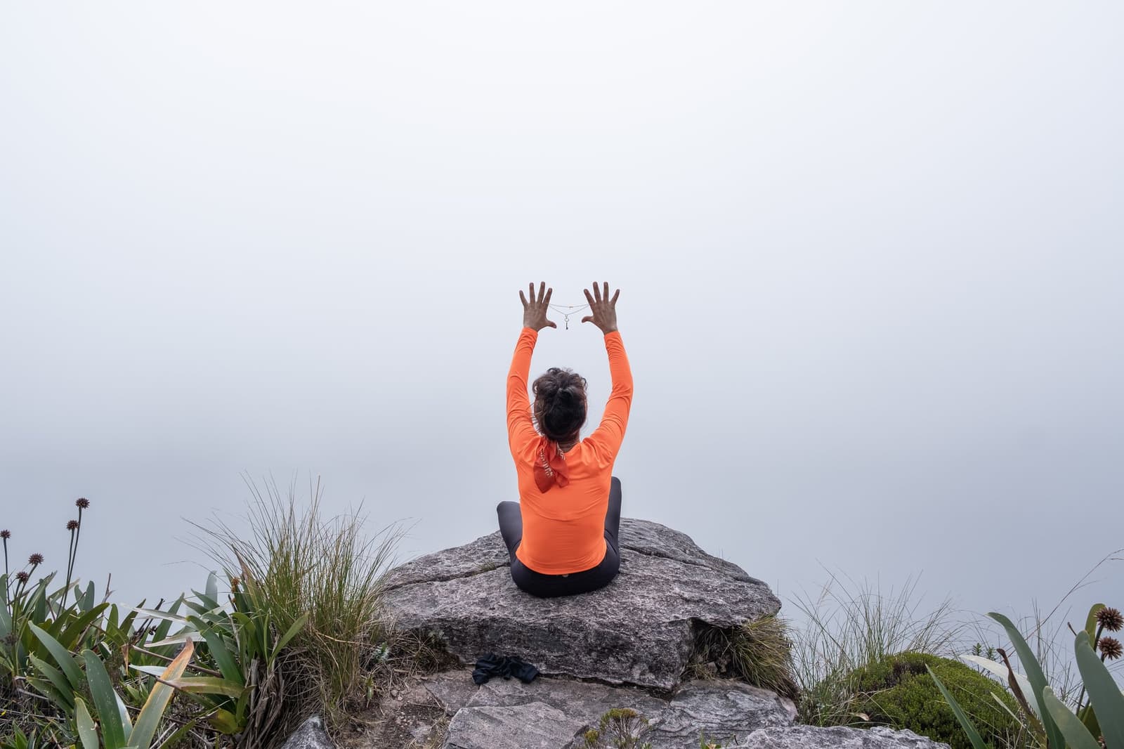 Mist tot aan de horizon, en toch voelt het als meer dan genoeg. Een persoon in een oranje shirt met lange mouwen zit met gekruiste benen op een rotsuitstulping op het plateau, armen omhoog boven het hoofd, omringd door hooglandvegetatie en dichte mist