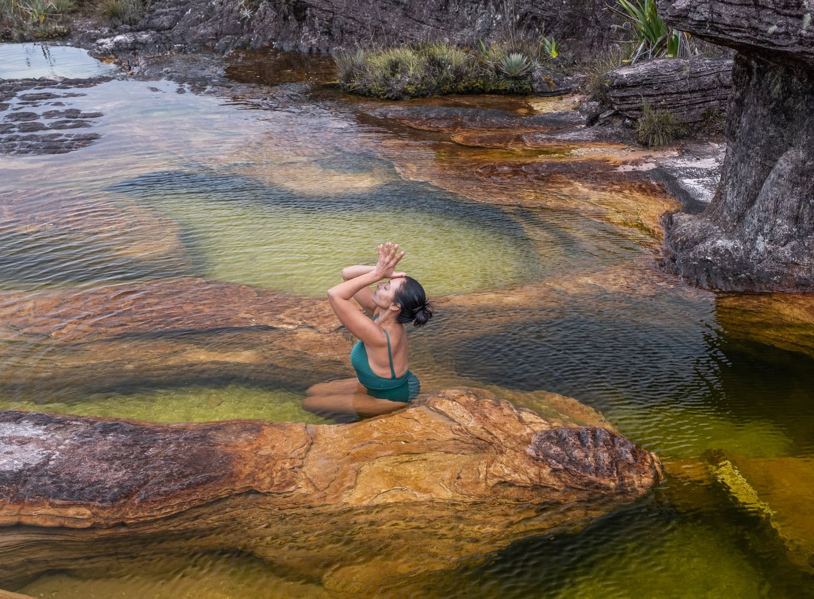 Ijskoud, glashelder, en precies wat je nodig hebt na een lange klim. Een vrouw in een groenblauw badpak knielt op een ijzerrijke oranje rotsplaat tijdens een Mount Roraima-expeditie, handen boven haar hoofd geheven, omringd door ondiepe heldere poelen met groengetint water en gelaagde oeroude steenformaties