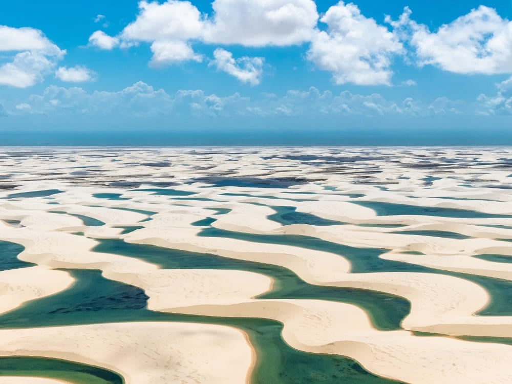 Photo: Julius Dadalti Veduta aerea di una vasta distesa di dune di sabbia bianca punteggiata da lagune stagionali blu e verdi, sotto un cielo azzurro con nuvole bianche, nel Parco Nazionale dei Lençóis Maranhenses, in Brasile.