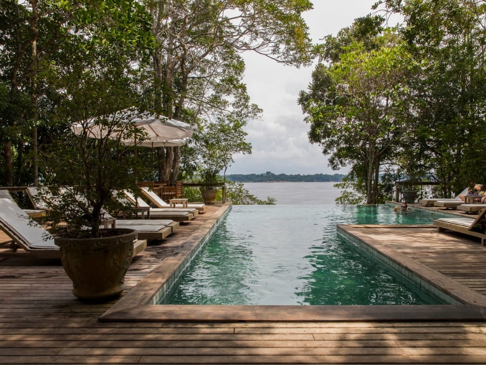 Piscina a sfioro su un deck di legno con lettini affacciati sul fiume, incorniciata dagli alberi della foresta in un ecolodge dell'Amazzonia.