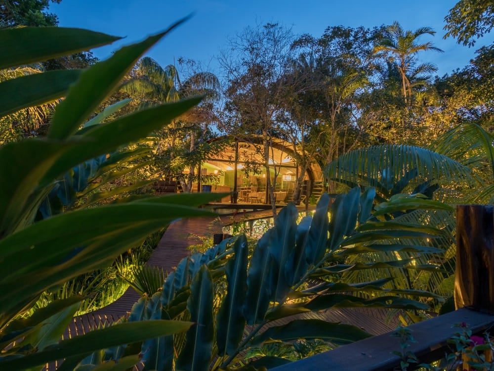Padiglione di un ecolodge dell'Amazzonia illuminato di luce calda, intravisto fra la vegetazione tropicale al crepuscolo.