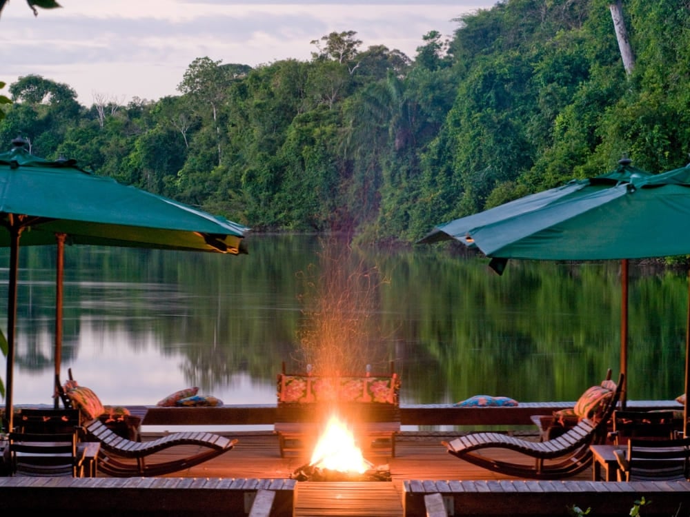 Deck di un lodge sul fiume al crepuscolo, con ombrelloni verdi, lettini e un braciere acceso riflessi nell'acqua calma del fiume amazzonico.