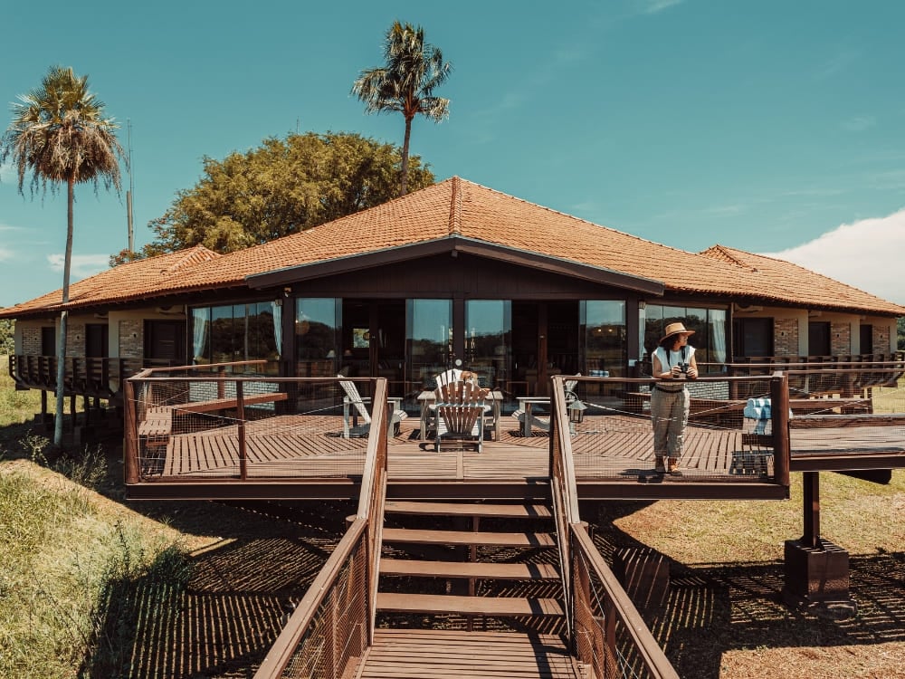 Photo: Felipe Castellari L'edificio principale di un lodge faunistico del Pantanal, con tetto di tegole e un ampio deck di legno sotto un cielo azzurro, e un ospite in piedi fra le palme.