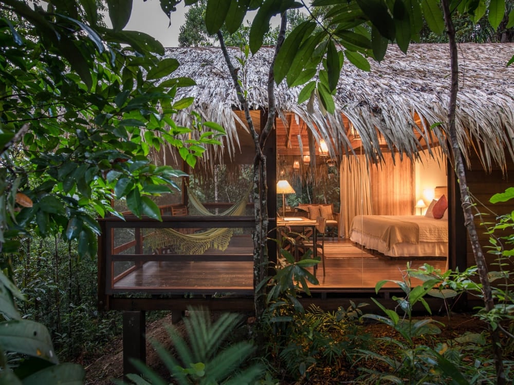 Bungalow di un lodge di lusso in Amazzonia, con tetto di paglia e pareti di vetro illuminate di luce calda al crepuscolo e un'amaca sul deck in mezzo alla foresta.