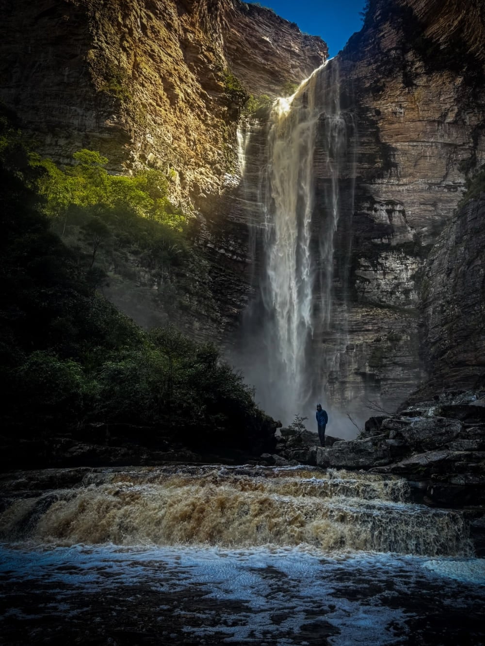 Un caminante solo en la base de la Cachoeira da Encantada, que cae entre las paredes del cañón sobre las aguas ámbar del Río Samina, en la Chapada Diamantina, Brasil. Un caminante solo en la base de la Cachoeira da Encantada, que cae entre las paredes del cañón sobre las aguas ámbar del Río Samina, en la Chapada Diamantina, Brasil.